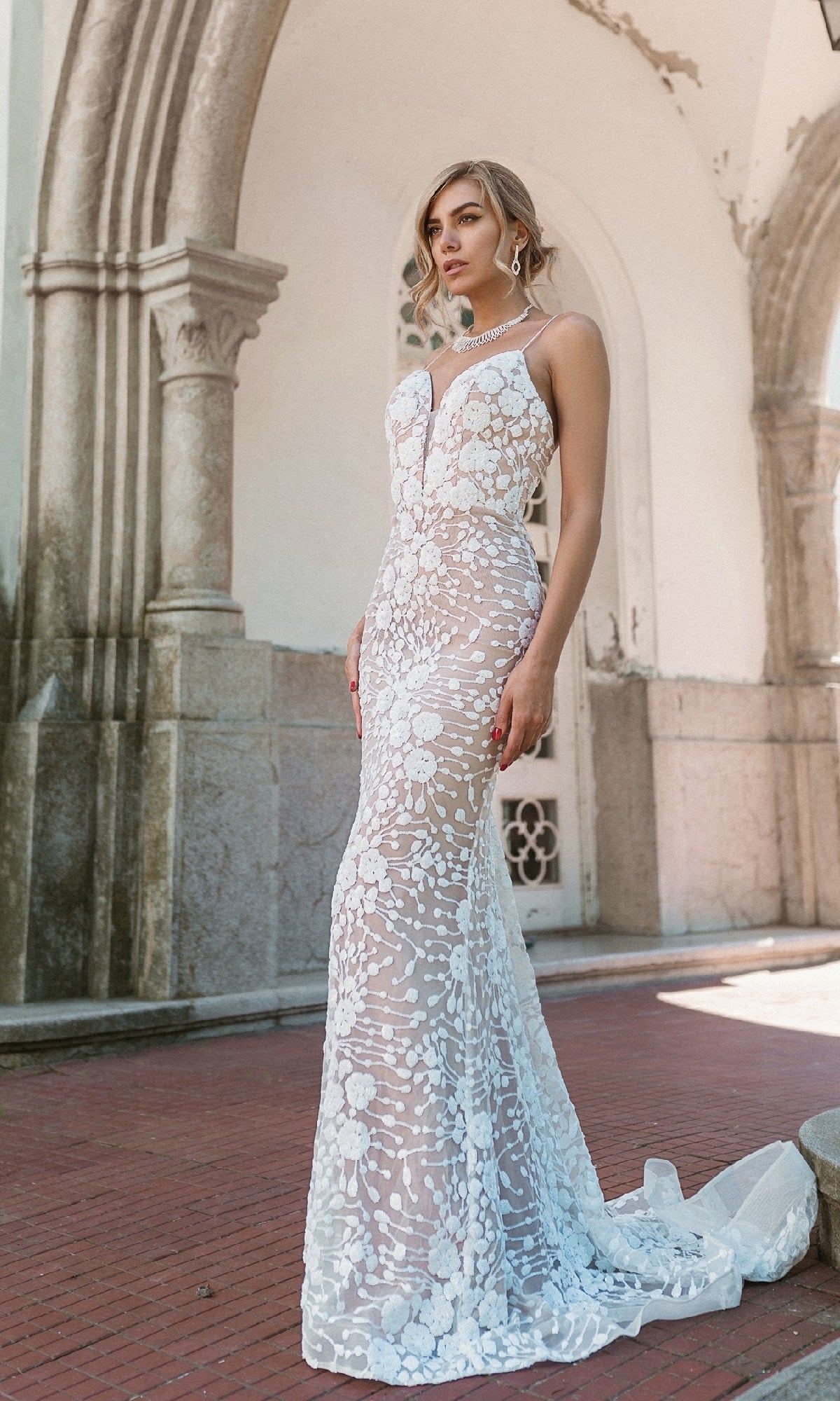 A woman poses outdoors in front of stone archways and a white building, wearing the Long White Lace Formal Dress: Tina Holly BB008—a fitted, sleeveless gown with floral lace patterns and a train.